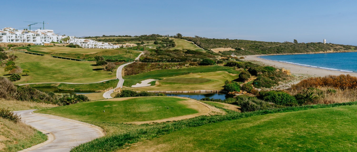 A golf course with a curving path leads towards the sea, with a beach on the right and residential buildings on the left under a blue sky.